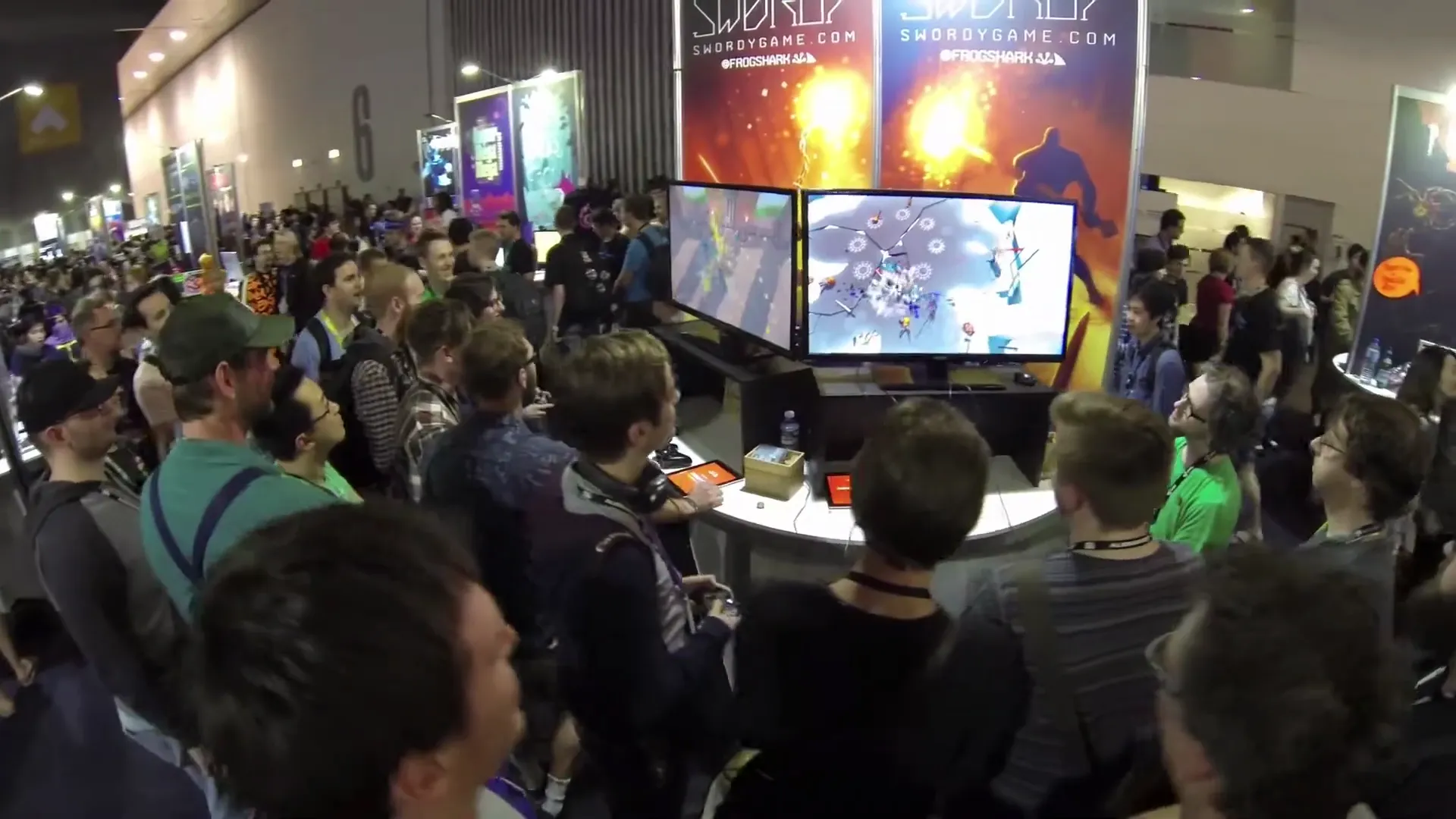 Wide view of a busy Swordy booth with players, spectators, screens, and banners.